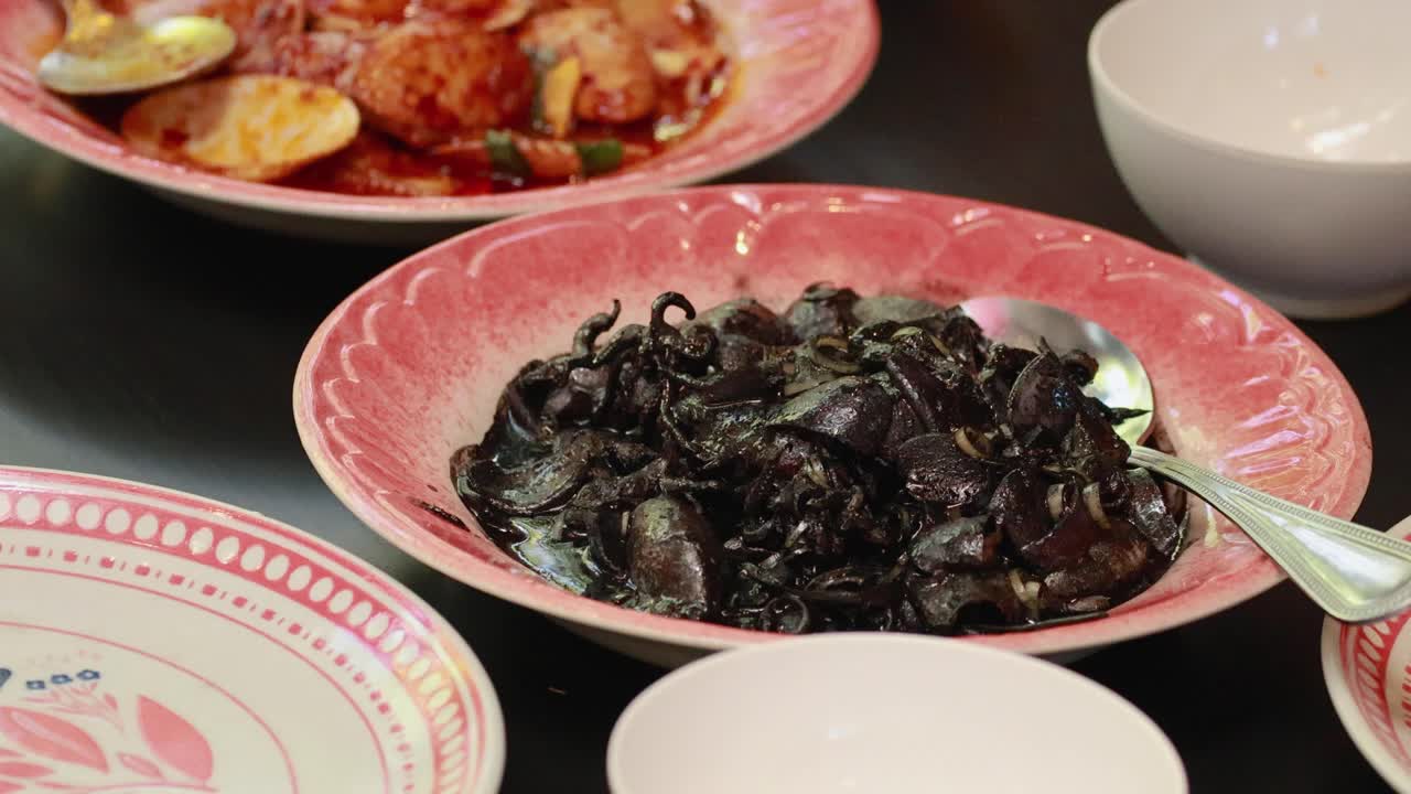 A person serves squid ink dish on a vibrant plate in a well-lit dining setting, highlighting seafood cuisine