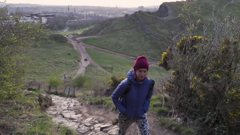 close up dolly slow motion shot of girl walking up the arthurs seat mountain on the hiking trail in evening during golden hour with city of edinburgh in the background down below