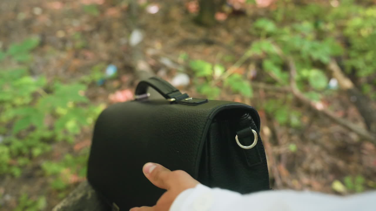 Close view of human hand placing jotter carefully inside black backpack and closing it while outdoors, showing detail of action with natural forest environment background symbolizing study