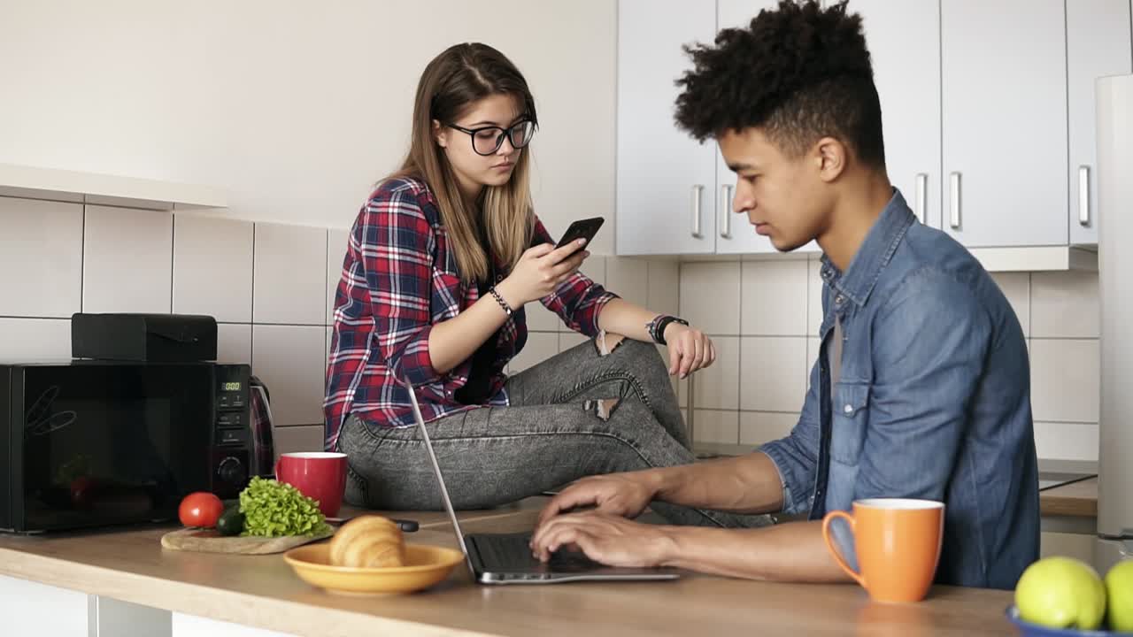 una joven pareja feliz de hipsters sentados en la cocina, ambos usando algunos dispositivos como portátiles, teléfonos inteligentes, sin hablar entre sí.
