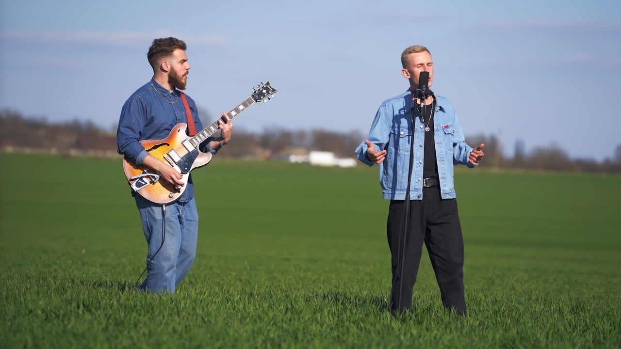 Two young men performing song in the green summer field. Blond man singing song at the microphone on stand under the electric guitar music.