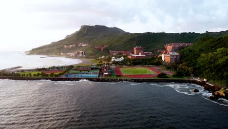 Ciudad-Costera-Al-Amanecer-Con-Exuberantes-Colinas,-Campos-Deportivos-Y-Un-Océano-Tranquilo,-Vista-Aérea