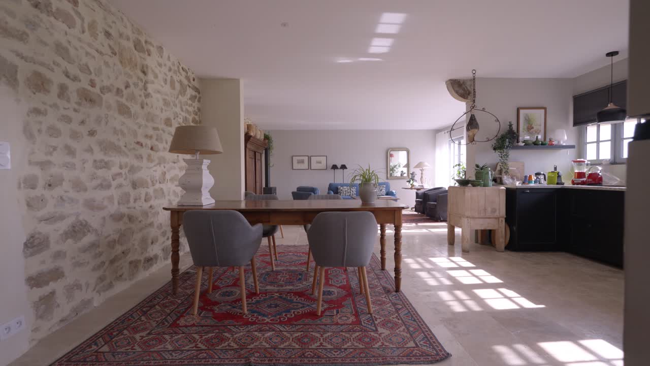interior of a property, large bright living room with dining room in foreground with wooden table and four gray chairs, light stone wall on left and kitchen on right, living room in background, carpet