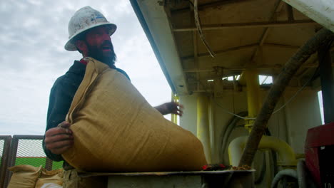 bearded man wearing dirty sweatshirt and protective helmet holding a full sack. man empties the bag with sawdust into the machine. low angle view.