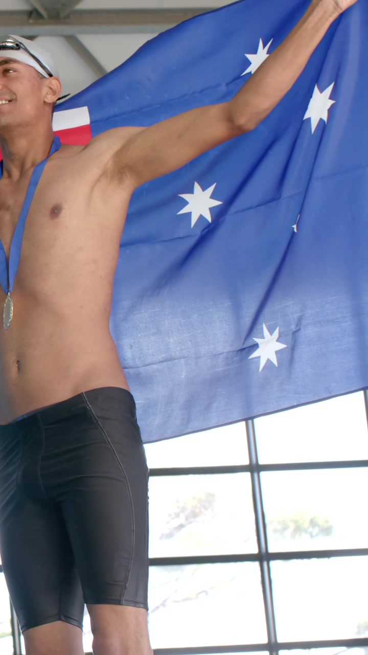 Video vertical: Nadador masculino sonriendo después de ganar una medalla, levantando una bandera azul con estrellas blancas junto a la ventana.