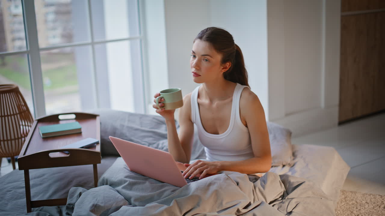 Primo piano di imprenditrice che lavora al computer a letto, godendosi il caffè del mattino sul materasso.