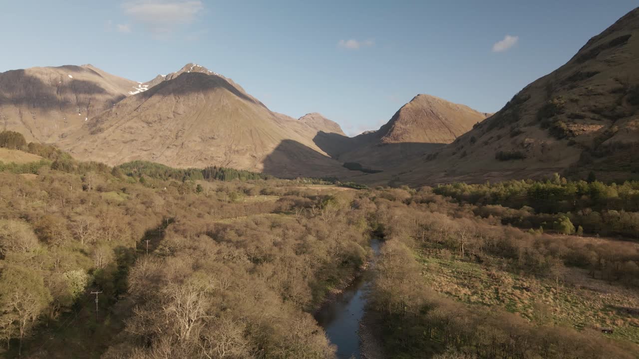 Drone flying along bottom of Scottish valley over trees and river on a summer day, featuring mountains in background