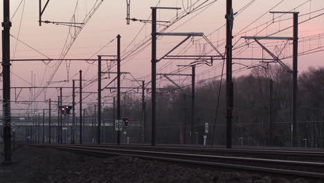 Vías-De-Tren-Y-Catenaria-Al-Atardecer-Con-Autos-Que-Pasan-Por-Un-Puente-Distante,-Creando-Una-Sensación-De-Soledad