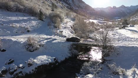 Seguimiento-Aéreo-De-Un-Coche-Cruzando-Un-Puente-De-Un-Río-En-Medio-De-Los-Pirineos-Españoles-Rodeado-De-Nieve.