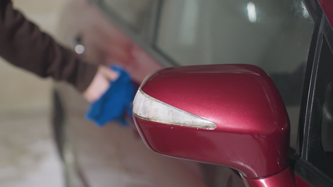 close up hand view of person wearing brown hoodie using blue cloth to clean shiny red car door near side mirror during detailing session inside garage under bright indoor lighting