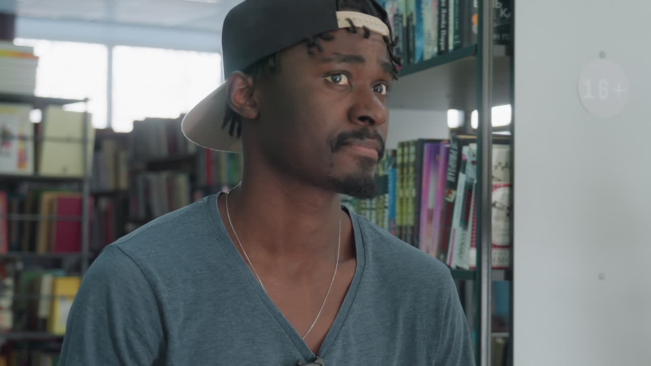 Side view of young man wearing casual shirt and backward cap standing near bookshelf in library, looking attentively at colorful row of books arranged neatly on shelves in well-lit reading area