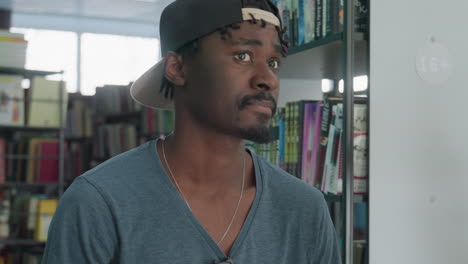 side view of young man wearing casual shirt and backward cap standing near bookshelf in library, looking attentively at colorful row of books arranged neatly on shelves in well-lit reading area