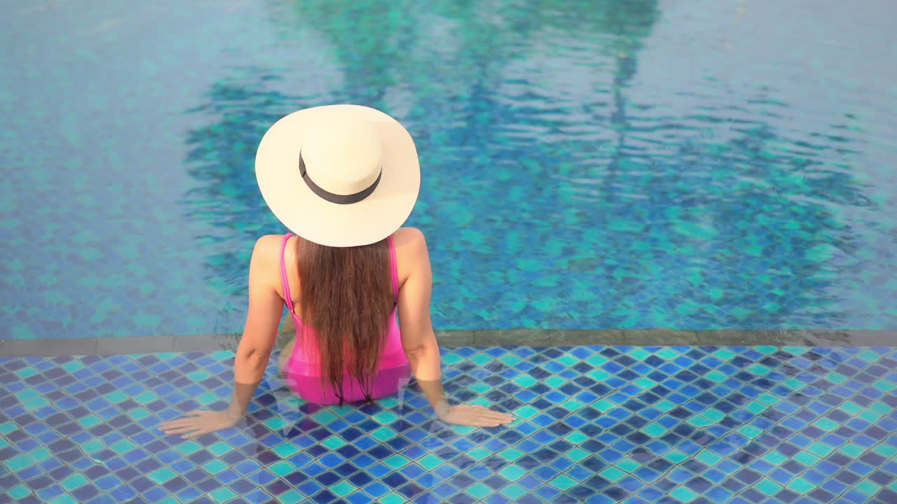 A woman with her back to the camera sits on the shallow step of a swimming pool