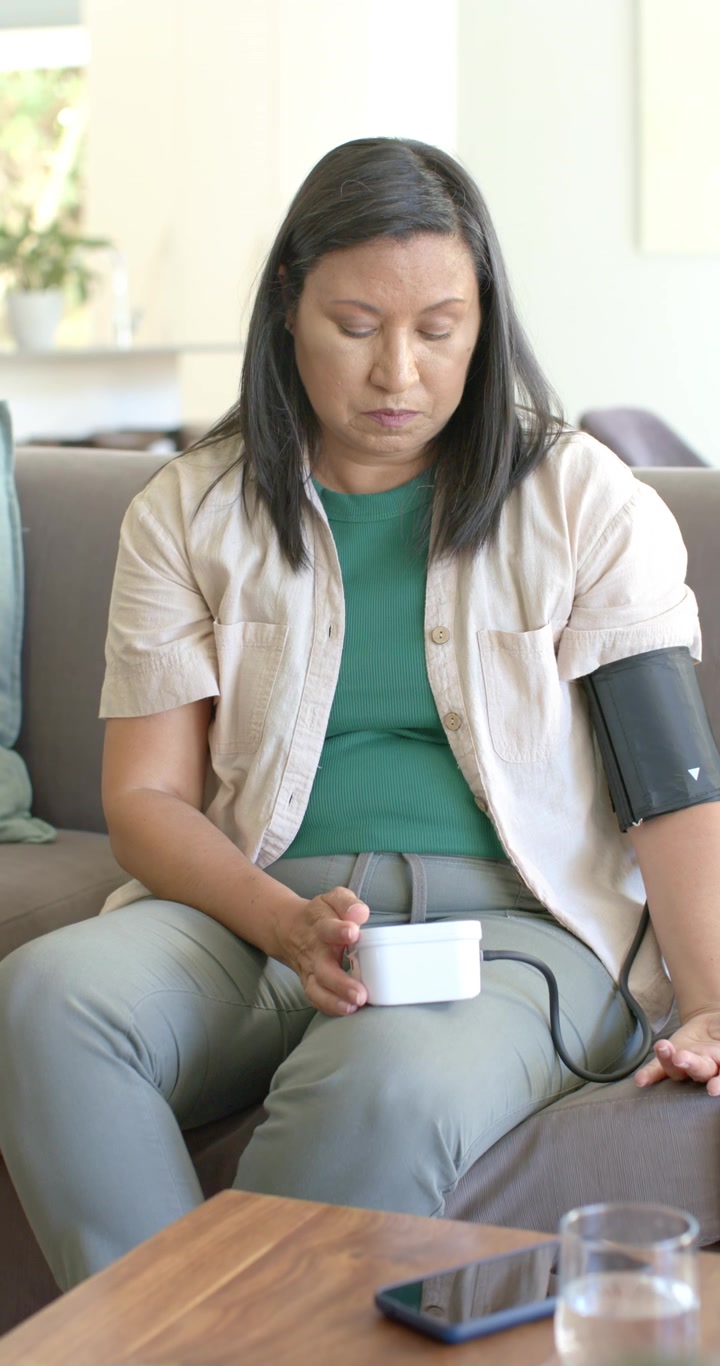 Video verticale: Donna adulta matura che usa un misuratore di pressione sul divano con il bracciale allacciato, in attesa del risultato.