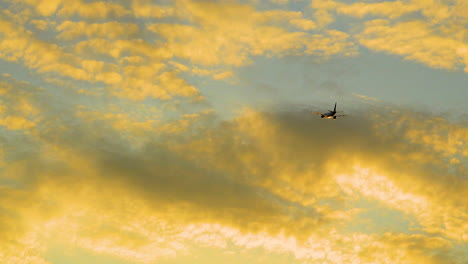 Jet-Airplane-Flying-Off-Into-Distance-Against-Golden-Hour-Sunset-Light-Being-Reflected-Off-Clouds
