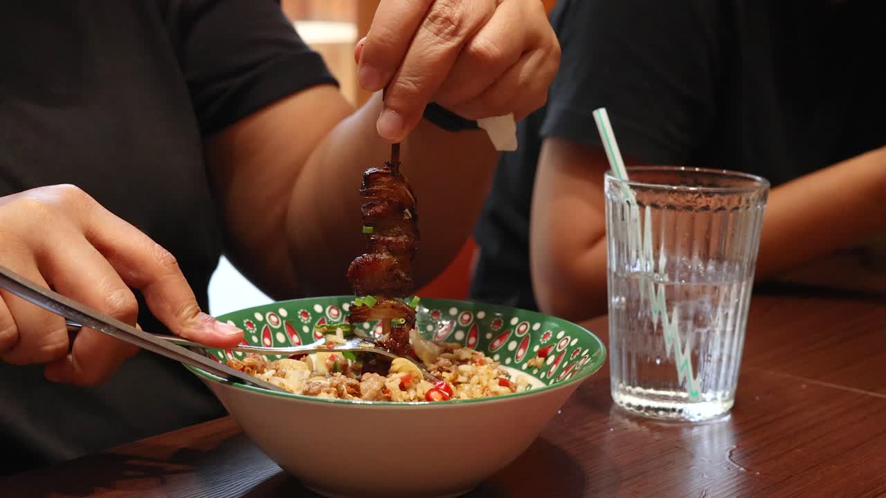 A person lifts a grilled beef skewer over a bowl of rice and vegetables in a casual restaurant setting, with natural lighting and a glass of water nearby
