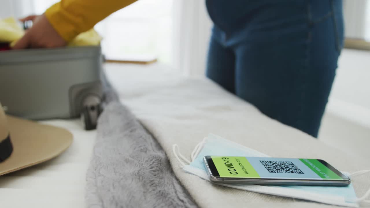 mujer afroamericana de talla grande empacando una maleta con pasaporte covid en un teléfono inteligente