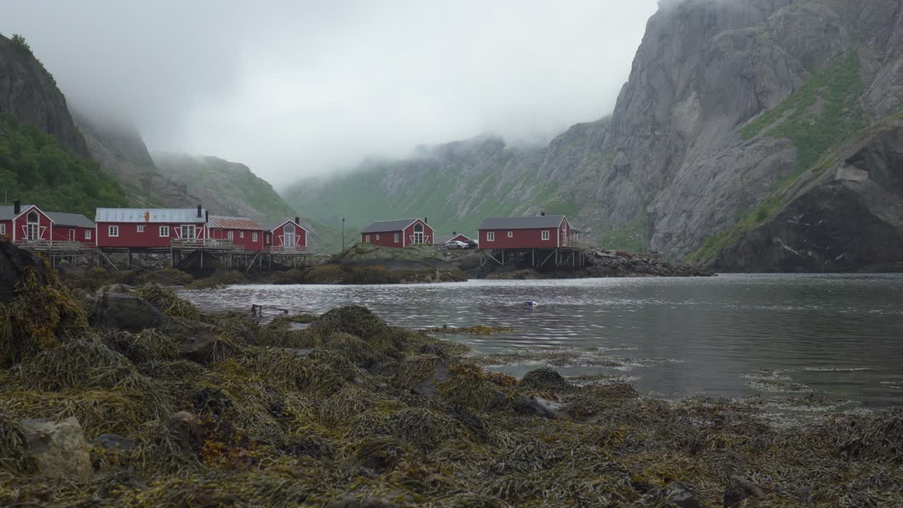 Lofoten, Norsko – Červené rybářské chaty v Nusfjordu se nacházejí podél klidného fjordu, obklopené strmými mlhavými horami – široký záběr