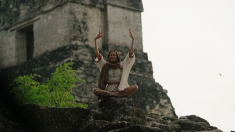 a woman sits cross-legged with hands together above her head, meditating in front of a mayan pyramid in tikal.