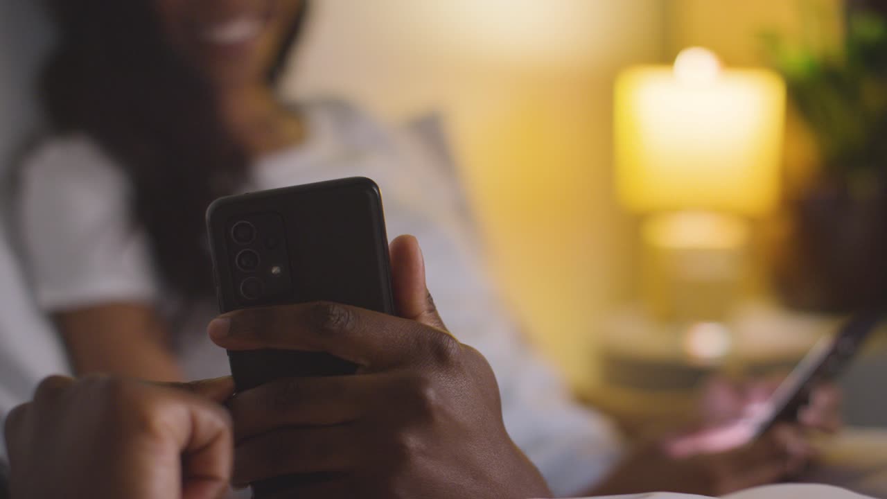close-up de una pareja sonriente en casa por la noche ambos mirando los teléfonos móviles en la cama
