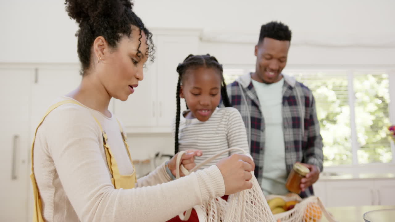 Happy african american parents and daughter unpacking shopping bag at home, slow motion