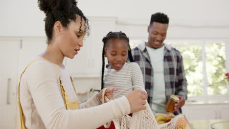 Glückliche-Afroamerikanische-Eltern-Und-Tochter-Packen-Zu-Hause-Ihre-Einkaufstasche-Aus,-Zeitlupe