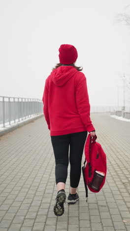 back view of athlete in red winter jacket and head warmer walking through fog-covered snowy landscape while carrying bag, surrounded by benches and trees blanketed in snow