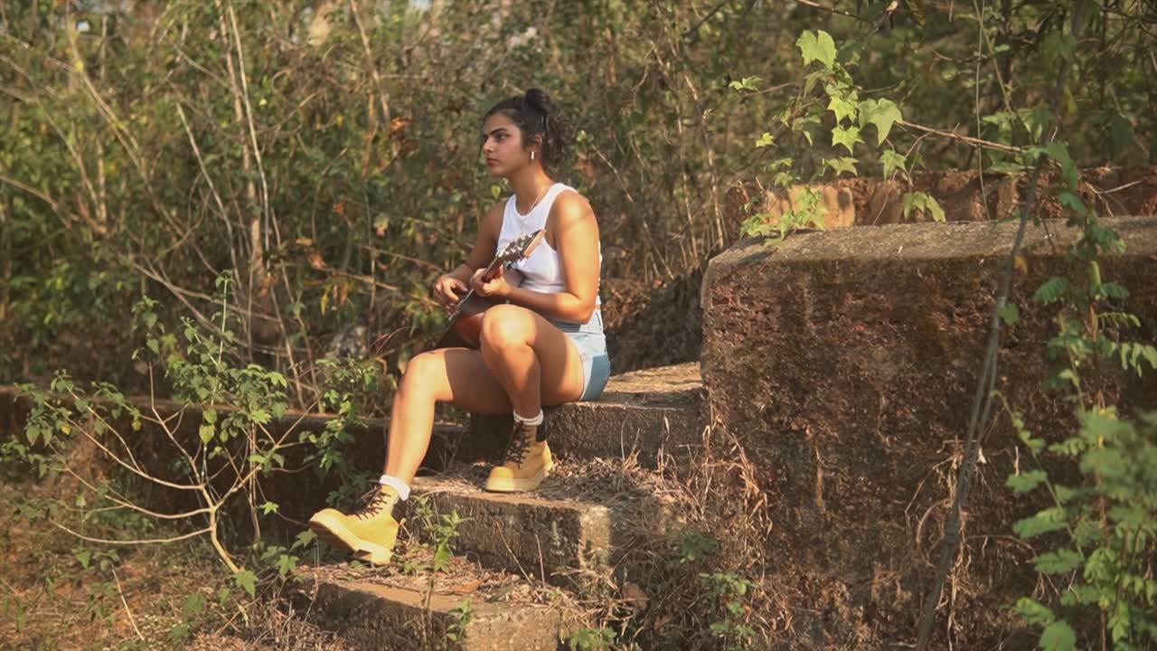 Donna indiana che suona la chitarra circondata dalla vegetazione durante il tramonto.