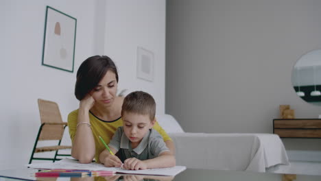 Mother-in-yellow-jacket-and-son-in-t-shirt-sit-at-the-table-and-draw-together-color-pencils-on-paper.-Happy-childhood.-Loving-helps-his-son-in-pre-school-training-and-develops-creativity-in-the-child