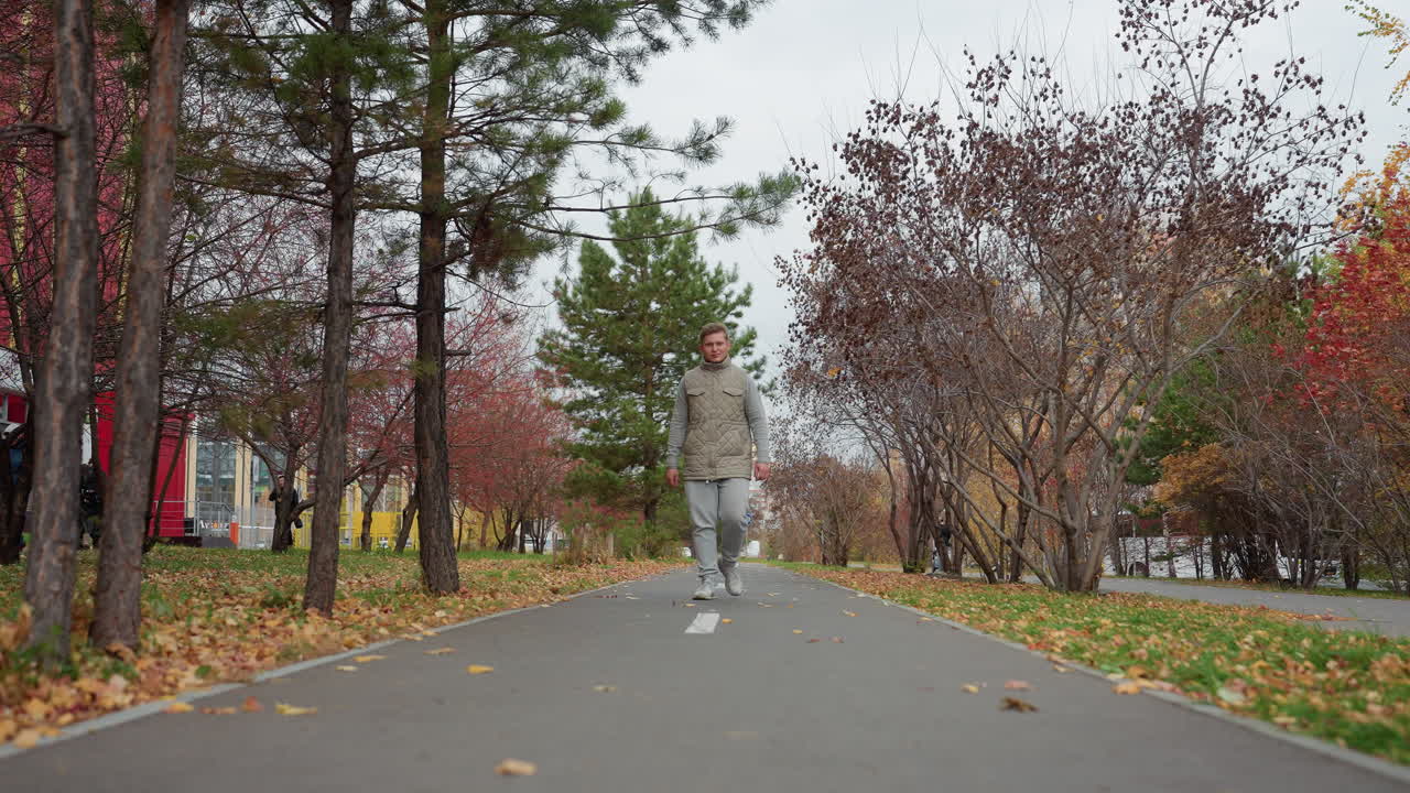 Confident young man walking on tarred path in park covered with scattered dry autumn leaves, surrounded by colorful trees, with people strolling in background on cloudy breezy day in urban setting
