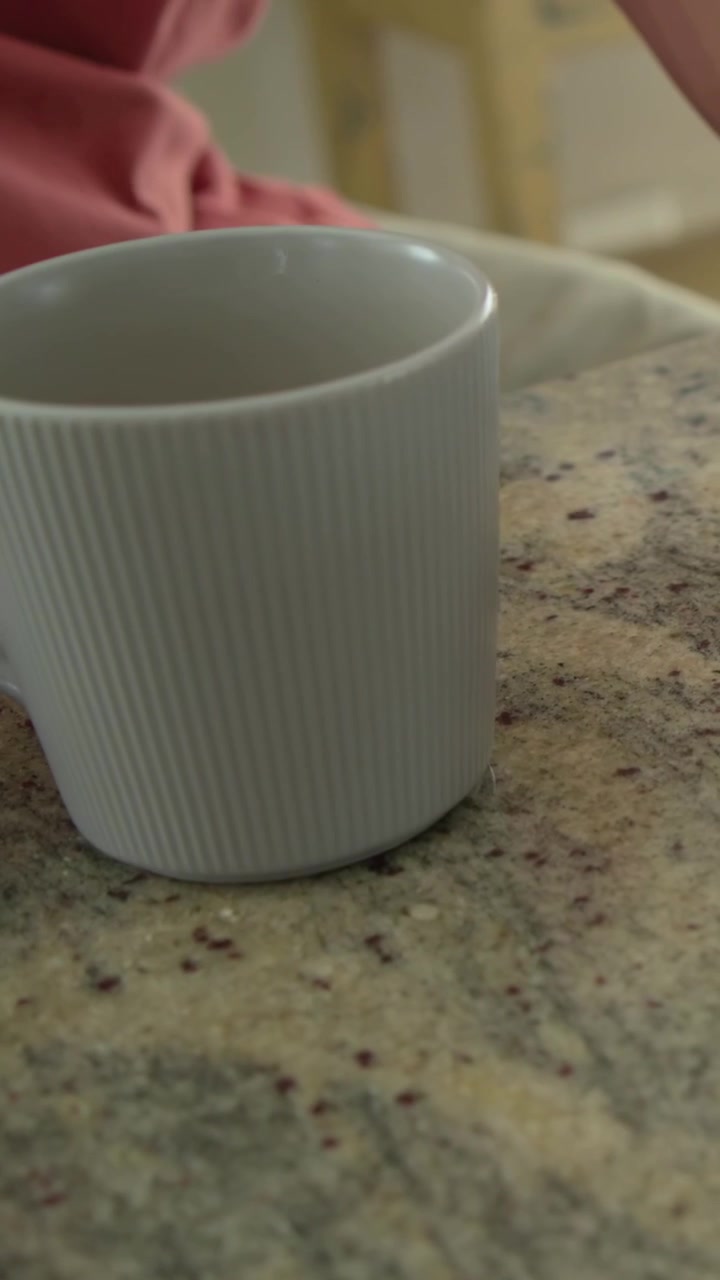 Vertical video: Arm entering frame woman in pink top reaching mug to warm hands placing on counter