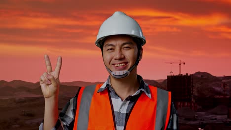 primer plano de un ingeniero asiático con casco de seguridad sonriendo y mostrando un gesto de paz mientras está de pie en el sitio de construcción, maquinaria de minería