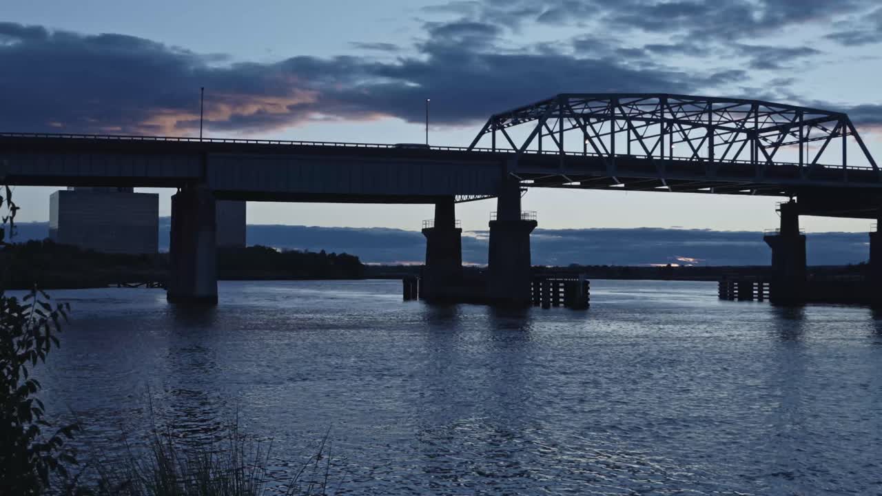 A semi-truck crosses a steel girder bridge at dusk, framed by a calm river below and a cloudy twilight sky above