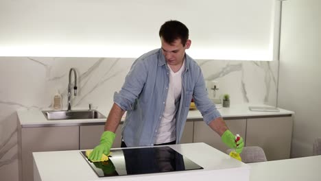 Hombre-Caucásico-Alto-Y-Alegre-Con-Camisa-Azul-Haciendo-La-Limpieza-En-La-Cocina,-Lavando-La-Estufa-De-La-Cocina-Con-Spray-Químico,-Interior-Moderno,-Limpieza-De-La-Casa.-Camara-Lenta