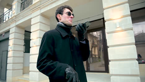 bottom view of a blind man in sunglasses and black coat outdoors while holding a walking stick and smartphone and having a hands free call