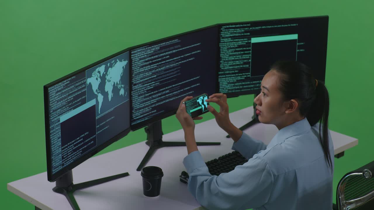 vista lateral de una desarrolladora asiática feliz mirando el código en un teléfono móvil mientras escribe código con múltiples pantallas de computadora en el estudio de pantalla verde