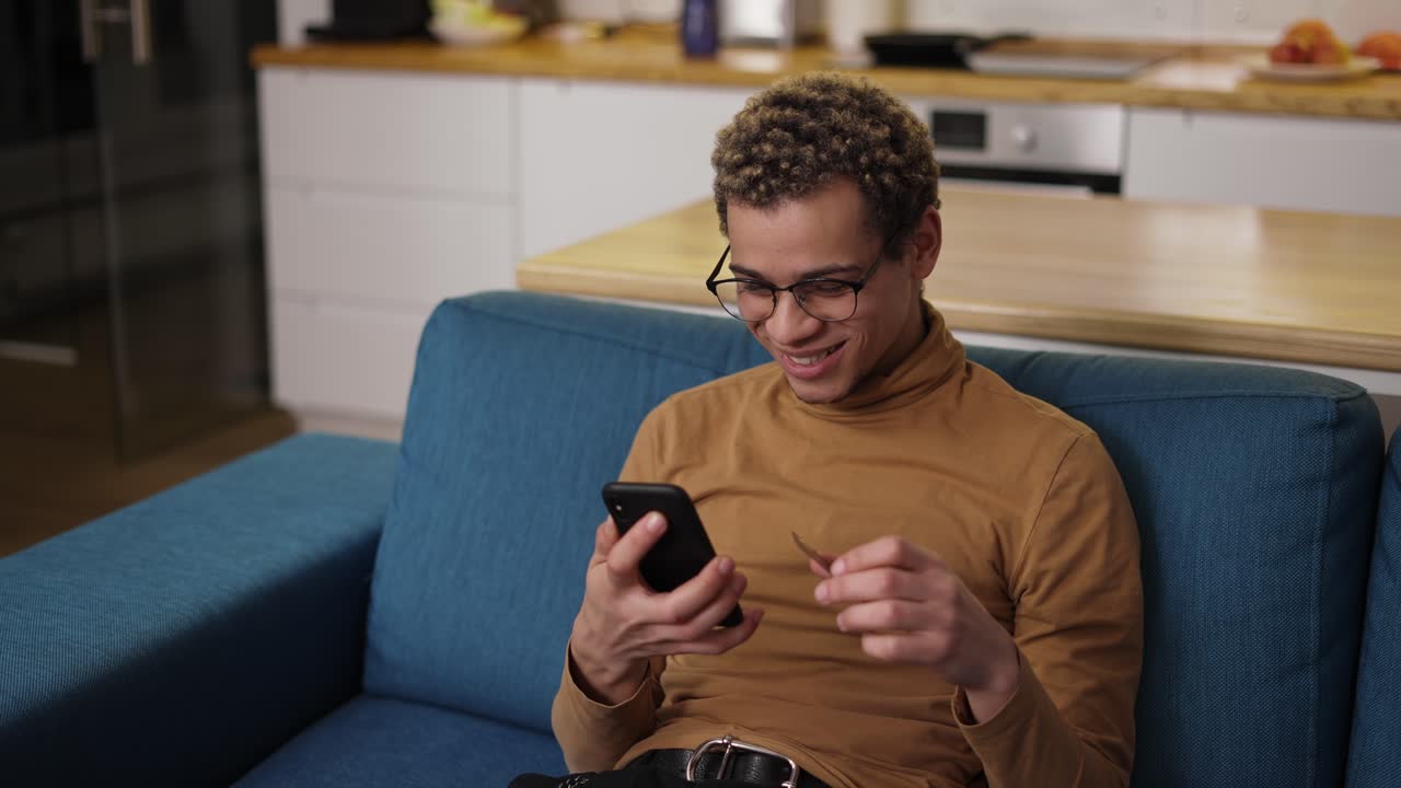 joven feliz comprando en línea usa un teléfono y una tarjeta de crédito en el sofá de casa