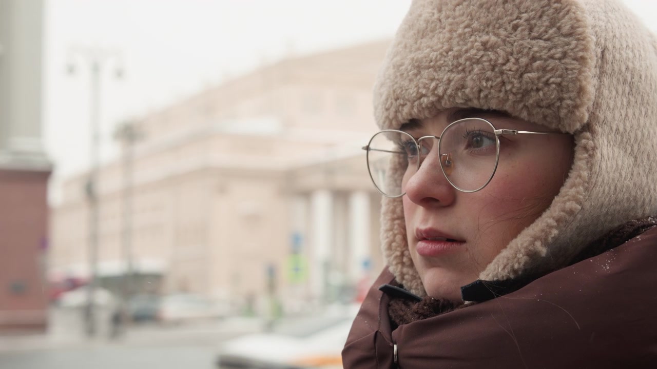 Tight medium close-up shows middle-aged person in thick knit trapper hat with fur trim, clear eyeglasses, and puffy coat, standing outdoors on cold overcast urban street with blurred buildings behind
