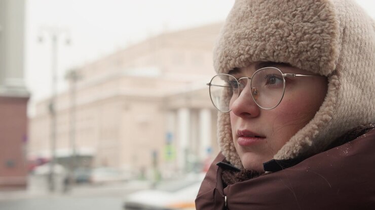 Primo piano medio ravvicinato di una persona di mezza età con un cappello da trapper in maglia spessa con bordo in pelliccia, occhiali trasparenti e un piumino, in piedi all'aperto su una fredda e nuvolosa strada urbana con edifici sfocati sullo sfondo.