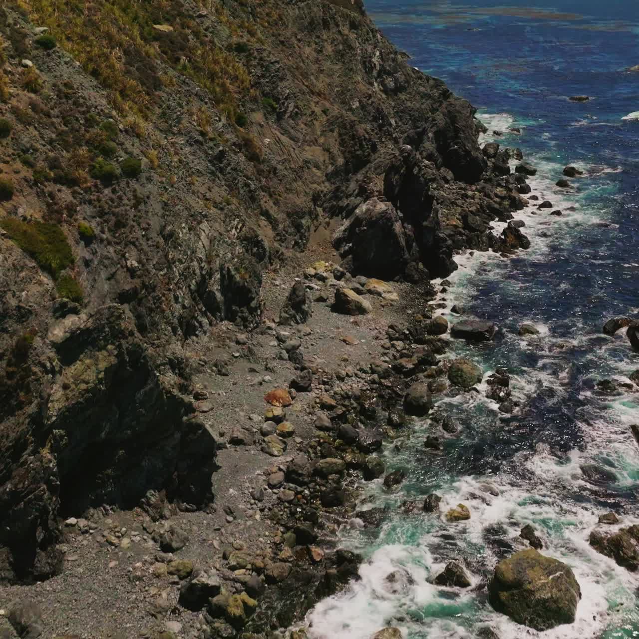 Flight over the rocky dangerous shore of California on sunny day. Foamy waves come to the coastline. Top view