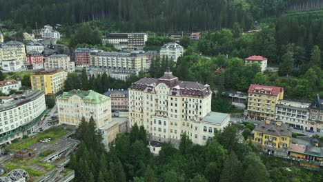 an aerial view of a european mountain town with a prominent historic grand hotel surrounded by colorful buildings and dense pine forests.