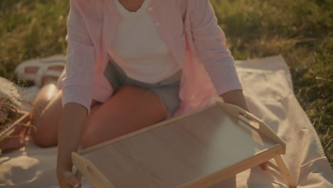 frau sitzt im freien auf einer matte und stellt einen picknicktisch mit blumen in einem gewebten korb neben sich unter sanftem sonnenlicht auf, um eine gemütliche picknickatmosphäre zu schaffen. sie ist in lässigen shorts und rosa hemd gekleidet