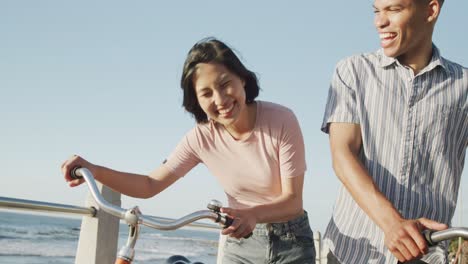 Happy-biracial-couple-walking-with-bikes-on-promenade,-in-slow-motion