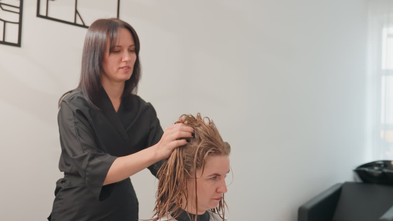 Salón de peluquería desenredando el cabello; estilista blanco cuidando las rastas de su cliente con peinado suave y separación por secciones. Interior moderno y tranquilo, luz natural suave, bata negra, expresión atenta, movimientos lentos y precisos.