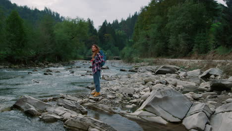 woman standing at bank of river in forest. girl hiking along mountain river