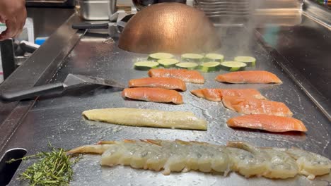 close-up of teppanyaki chef’s hands adding water to daikon radish and covering to steam. zucchini, sizzling rosemary and thyme, salmon, shrimp skewers, and white fish also cooking on the grill.