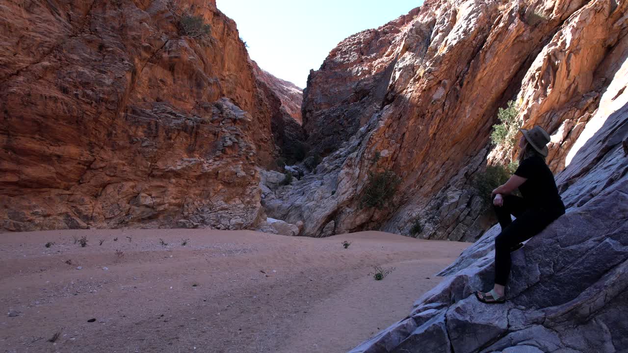 Drone flies past a person seated on a rock in a narrow canyon with sandy ground, capturing intimate outdoor moments framed by rugged terrain