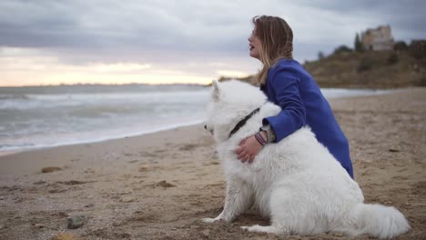 Side-view-of-a-young-woman-sitting-on-the-sand-and-embracing-her-dogs-of-the-Samoyed-breed-by-the-sea.-One-of-the-starts-to-run