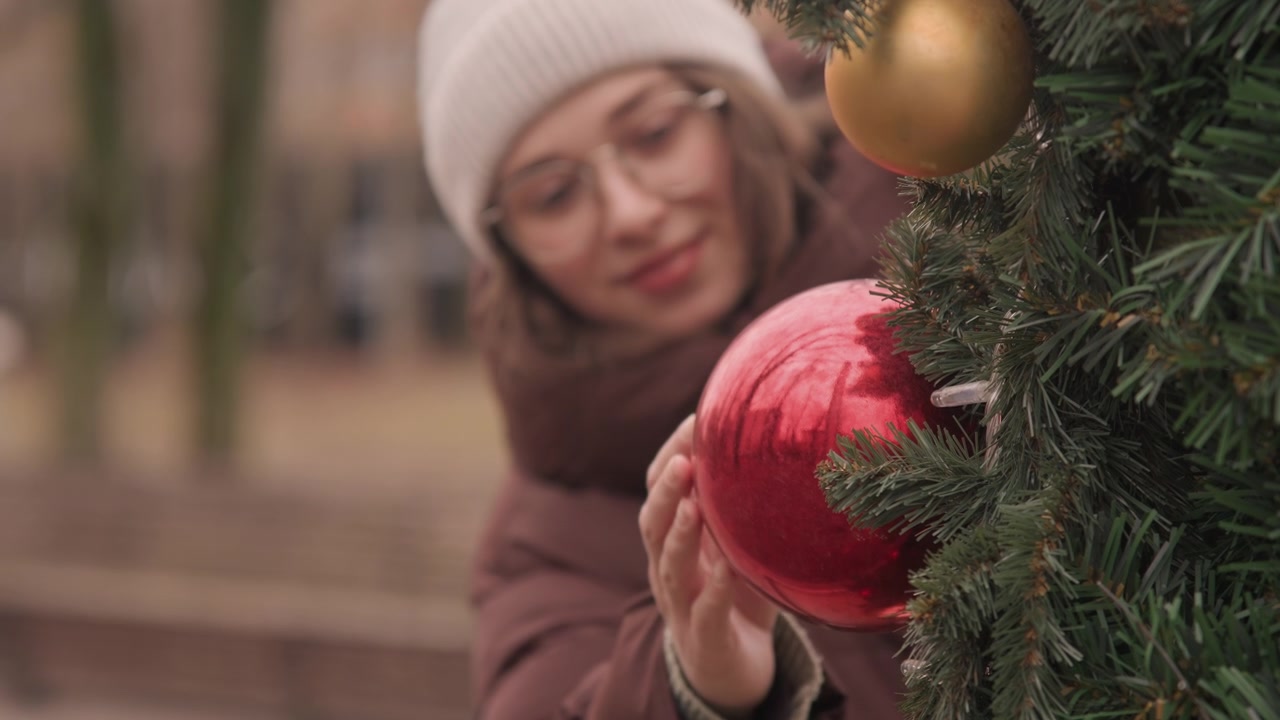 Jovem com gorro descobre uma bola vermelha em uma árvore de Natal ao ar livre, estudante fazendo uma pausa em um banco de parque, toque suave na decoração brilhante, luz suave de inverno e fundo urbano desfocado
