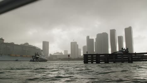 cruise ship docked in a large port in a city, with yachts and boats moving and coming into view under a cloudy sky